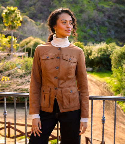 A woman wearing a brown cropped jacket with flap pockets over a white turtleneck and black pants, standing outdoors on a balcony with a green hillside background.