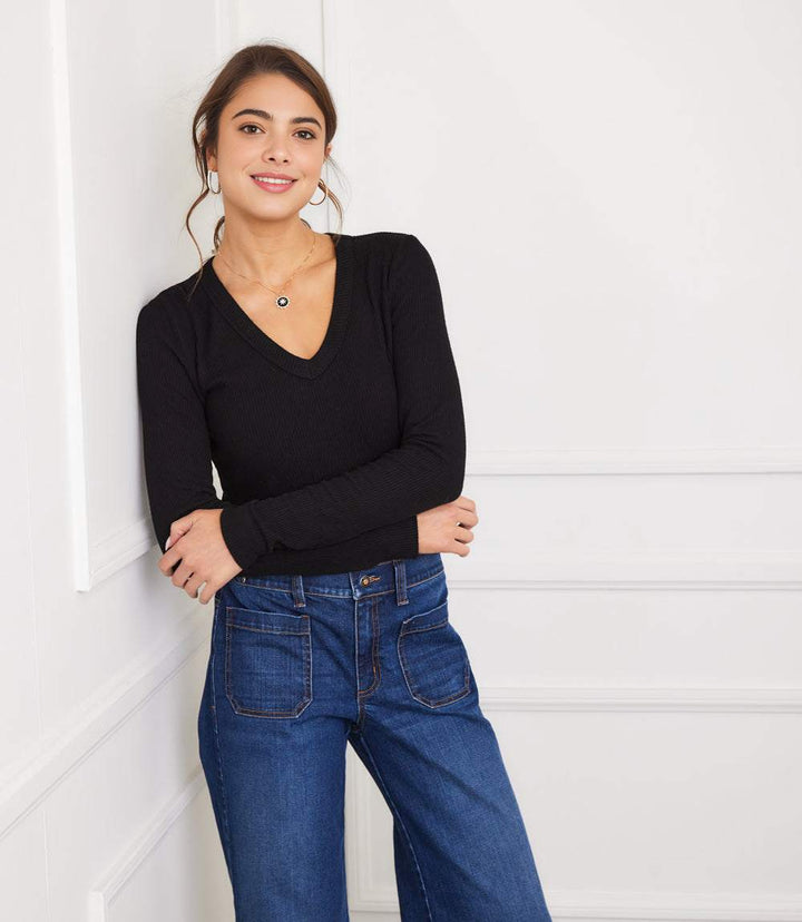 Woman wearing a black long sleeve ribbed V-neck top with slim fit and blue jeans, posing against a white wall.