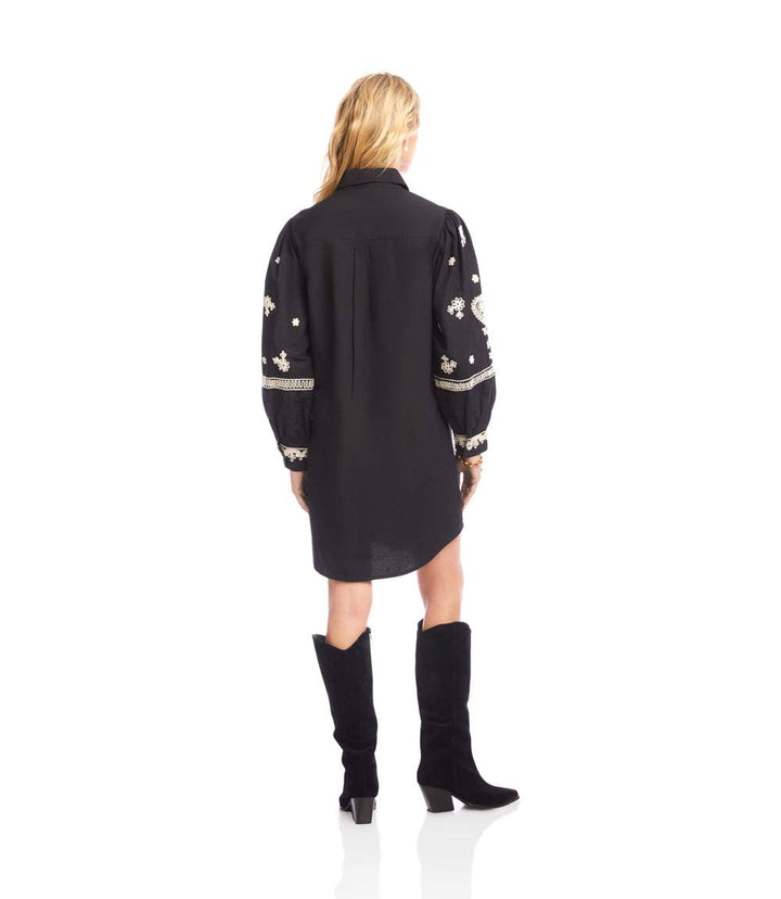 Back view of a woman wearing a black blouson sleeve shirtdress with cream rope-stitch embroidery and tall black boots