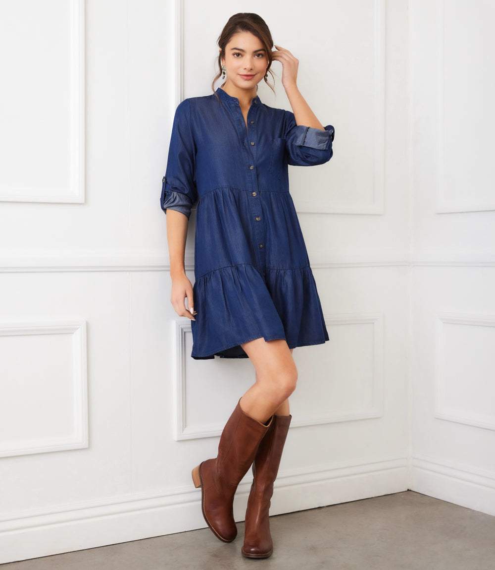 Woman wearing a navy blue roll tab tiered shirtdress with button front and brown boots in a white room