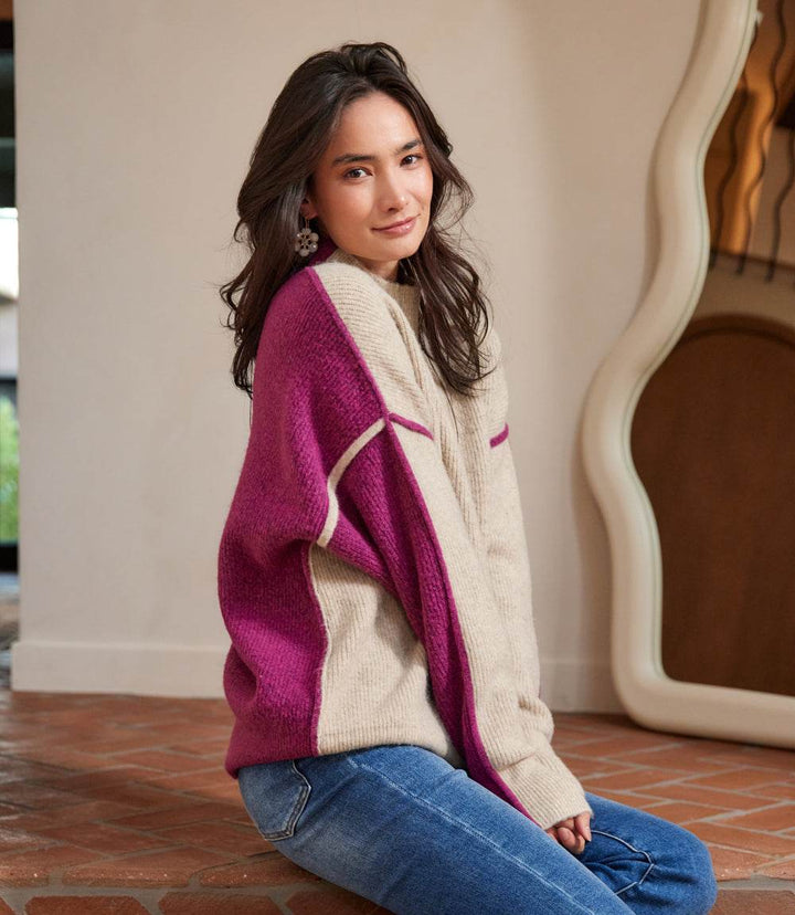 Woman wearing petite size colorblock mock neck sweater with purple and beige panels and drop shoulder sleeves sitting indoors