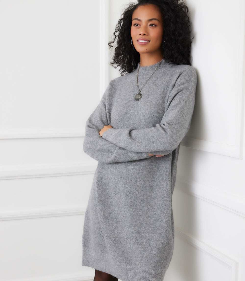 Woman wearing grey mock neck sweater dress with long sleeves and ribbed cuffs standing in white room