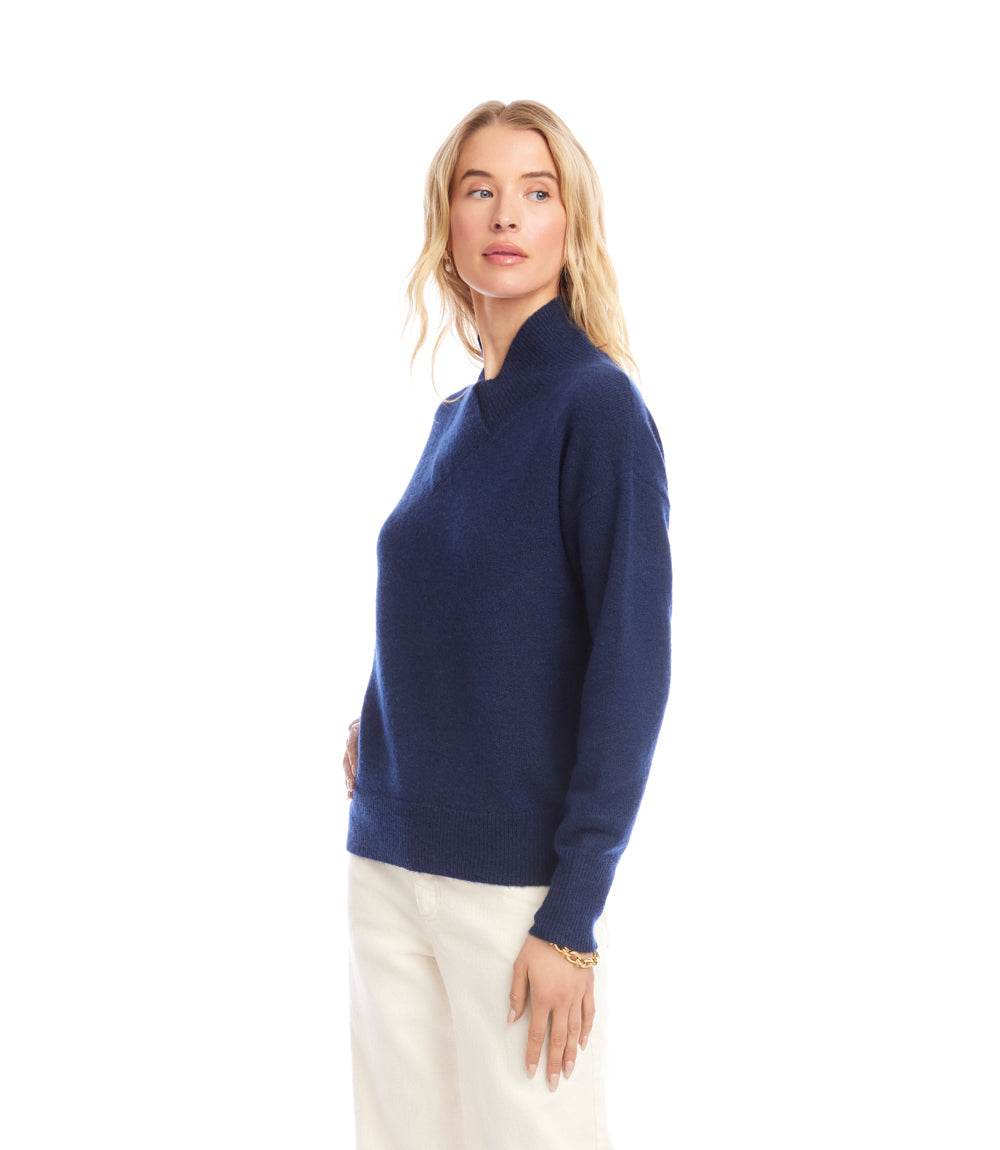 Woman wearing a navy shawl collar sweater with drop shoulder sleeves and ribbed cuffs, paired with white pants.