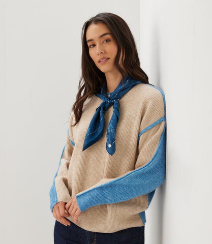 A woman wearing a beige ribbed knit sweater with bright blue color-blocked sleeves and a matching blue neck scarf, standing against a plain white wall.
