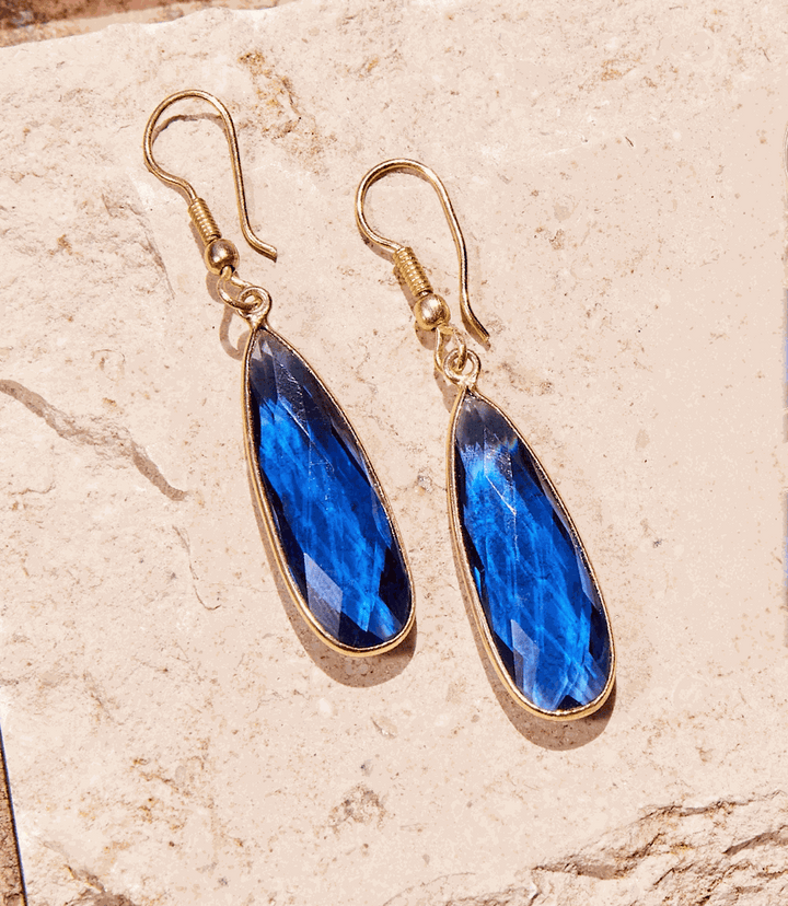 Pair of gold-tone dangle blue quartz earrings with faceted teardrop stones on natural stone background