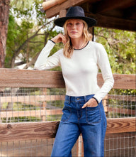 Woman wearing white long sleeve edge stitch crewneck top with black hat and blue jeans outdoors