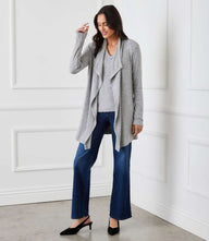 Woman wearing a grey ribbed drape front cardigan with blue jeans and black heels in a white room