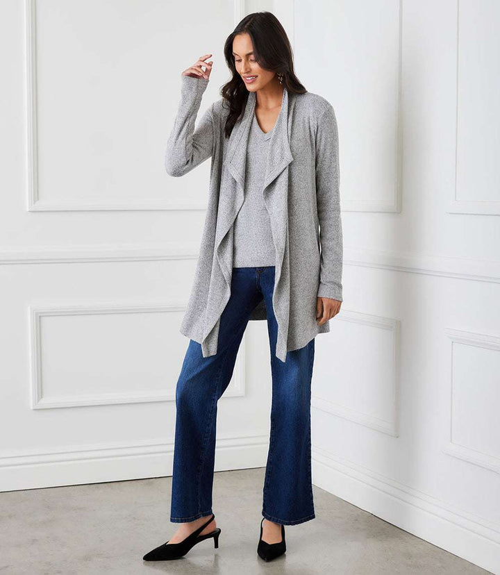 Woman wearing a grey ribbed drape front cardigan with blue jeans and black heels in a white room