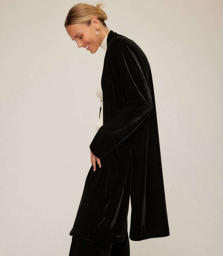 Black stretch velvet duster coat with open front, long sleeves, side slits, and front pockets worn by woman
