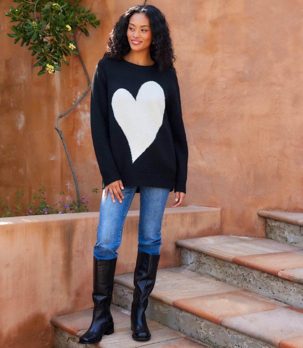 Woman wearing black Heart Jacquard Sweater with white heart motif, paired with jeans and black boots.