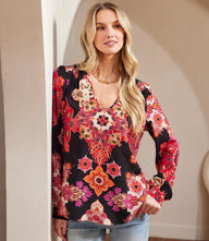 Woman wearing long sleeve shirred blouse with red, magenta, and cream floral print on black background and u-neckline.