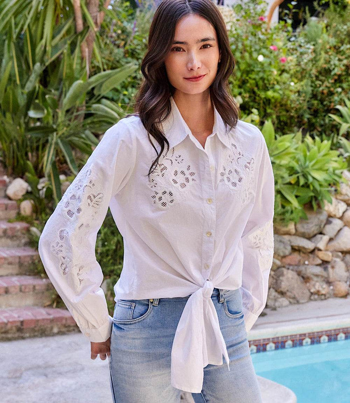 Woman wearing white embroidered tie front top with eyelet floral design and button closure by poolside