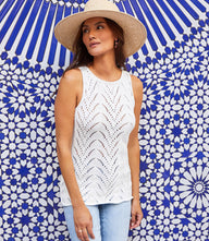 Woman wearing white sleeveless crochet sweater tank with scoop neck and light blue jeans against blue patterned background