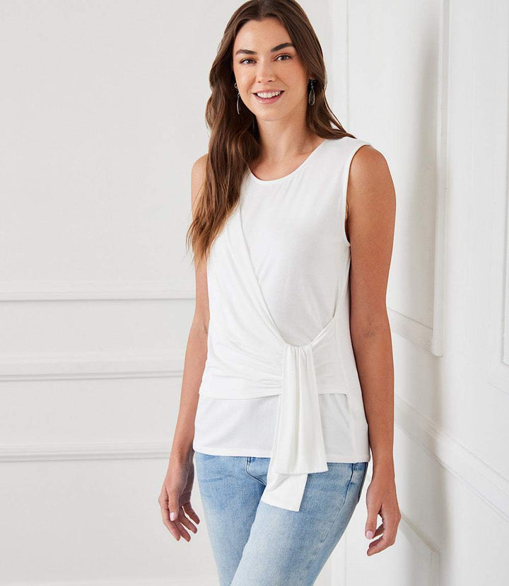 Woman wearing a white sleeveless side drape top with a scoop neck and side-tie detail paired with light blue jeans