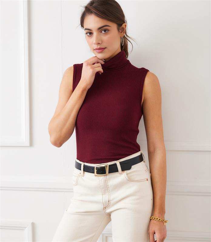 Woman wearing sleeveless ribbed turtleneck tank in burgundy paired with white pants and black belt