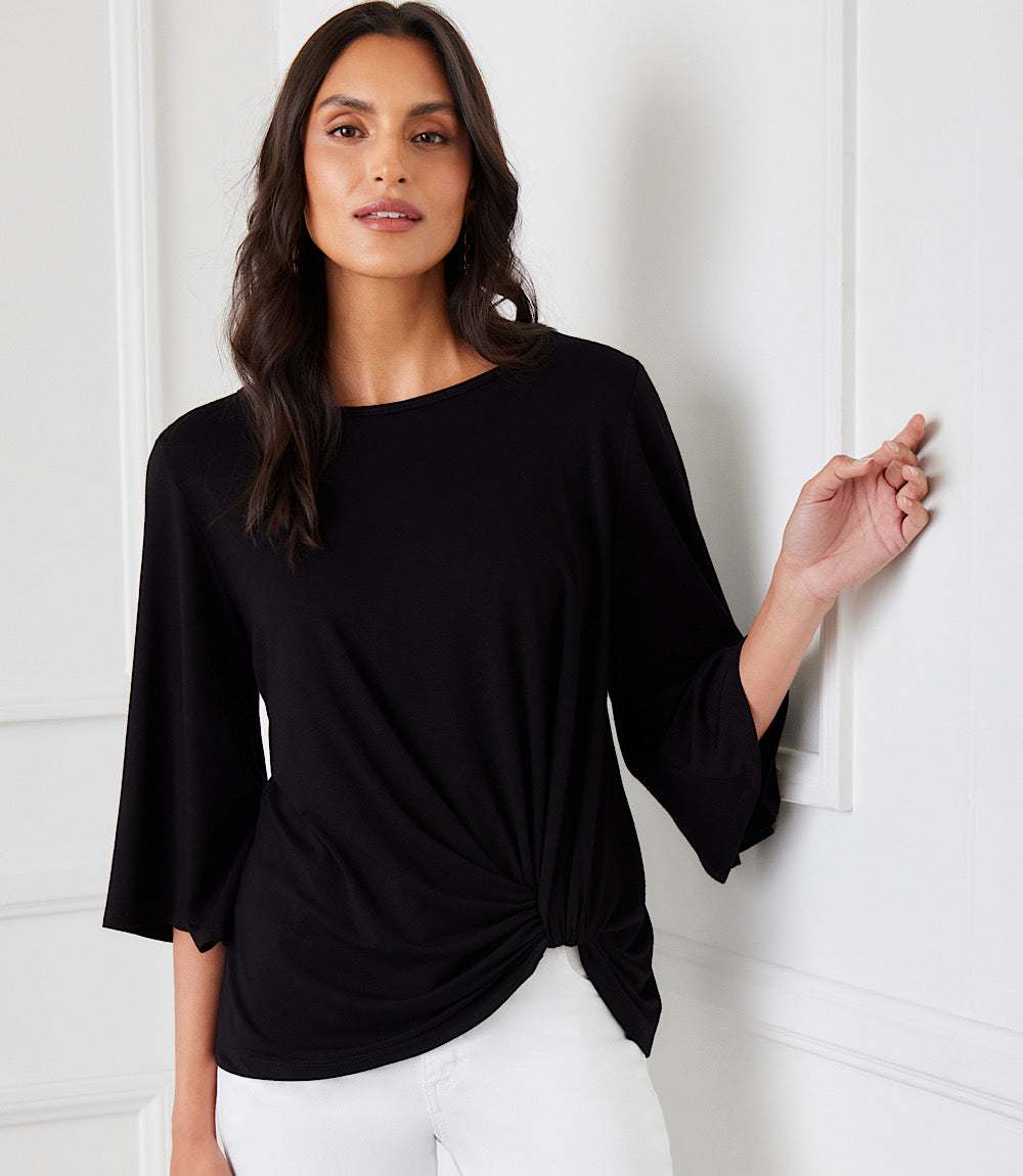Woman wearing black flare sleeve pick-up top with boat neck and twist hem detail paired with white pants in elegant indoor setting