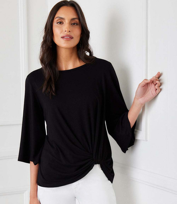Woman wearing black flare sleeve pick-up top with boat neck and twist hem detail paired with white pants in elegant indoor setting