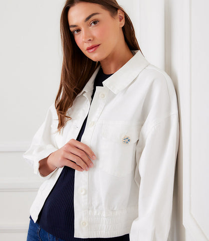 Woman wearing a white jacket with a small decorative item, standing against a white wall.