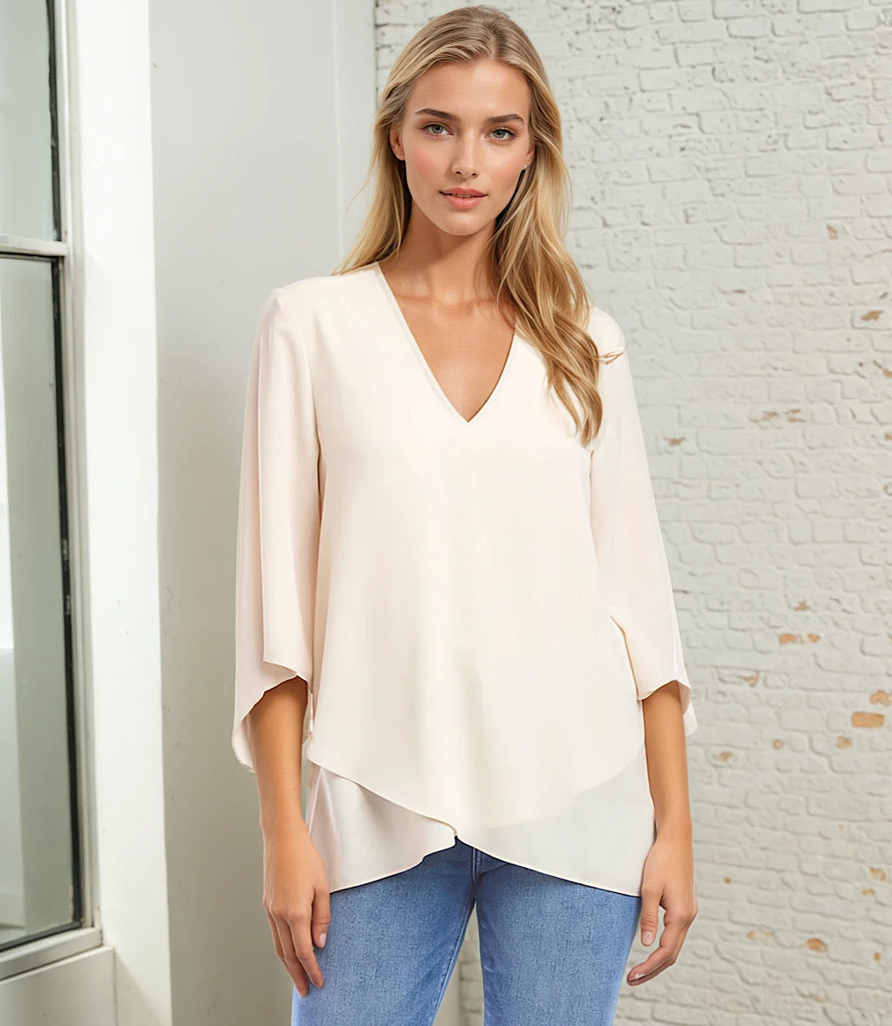 Woman wearing a light pink Bracelet Sleeve Asymmetric Top with V-neck, flare sleeves, and layered asymmetrical hem paired with blue jeans in a bright room