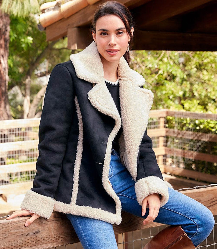 Woman wearing black faux suede shearling jacket with cream shearling trim sitting outdoors