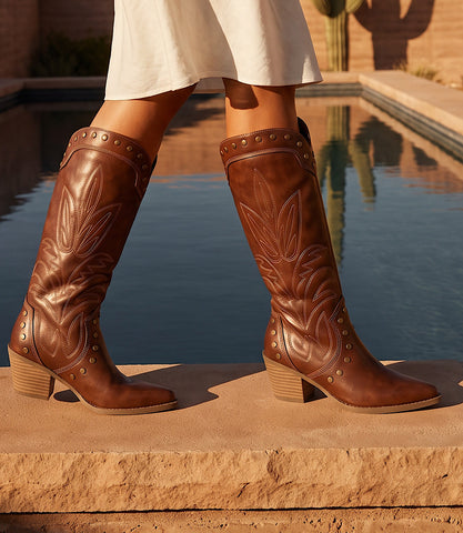 A person wearing knee-high brown leather cowboy boots with carved western detailing and stacked block heels, standing on a stone ledge beside a pool.