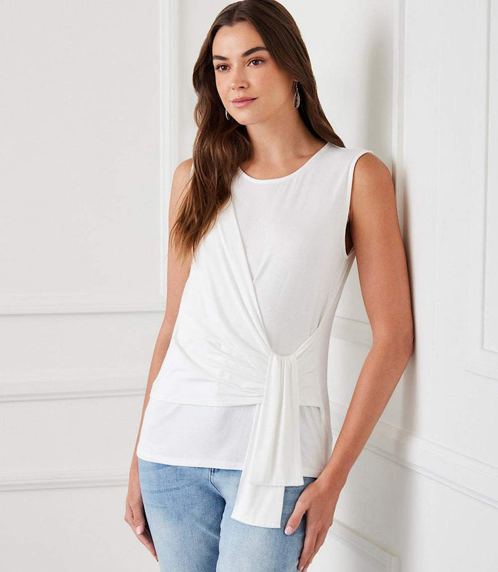 Woman wearing white sleeveless side drape top with scoop neck and side-tie detail paired with blue jeans