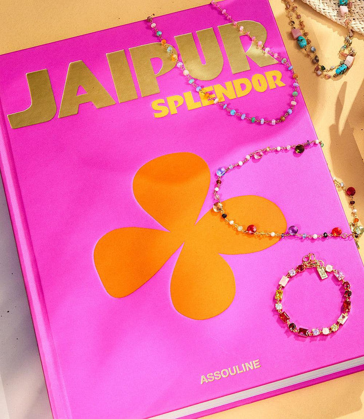 Bright pink hardcover book titled Jaipur Splendor with gold lettering and an orange flower design, placed alongside colorful beaded jewelry.