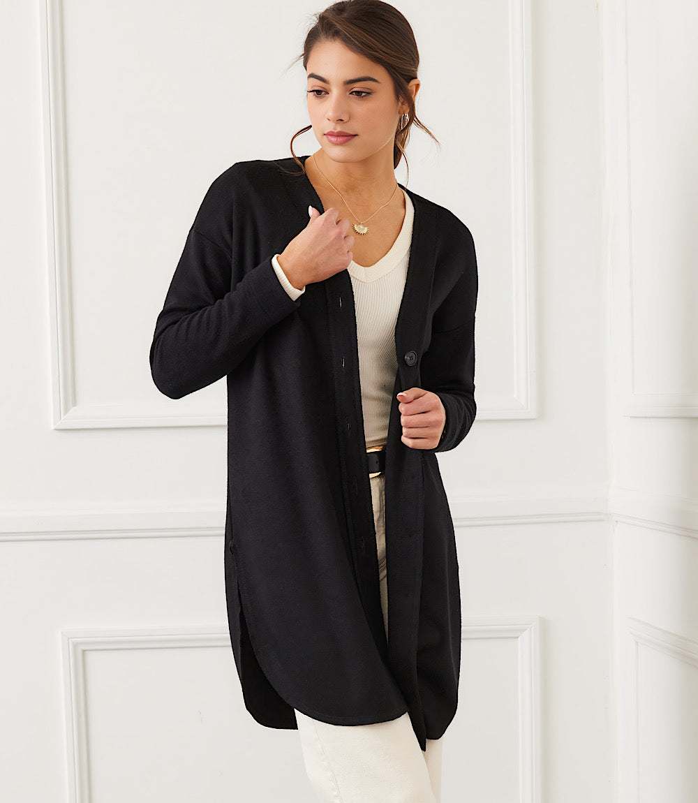 Woman wearing a black long sleeve shirttail cardigan with a V-neck and button front closure, styled with white pants and a beige top.