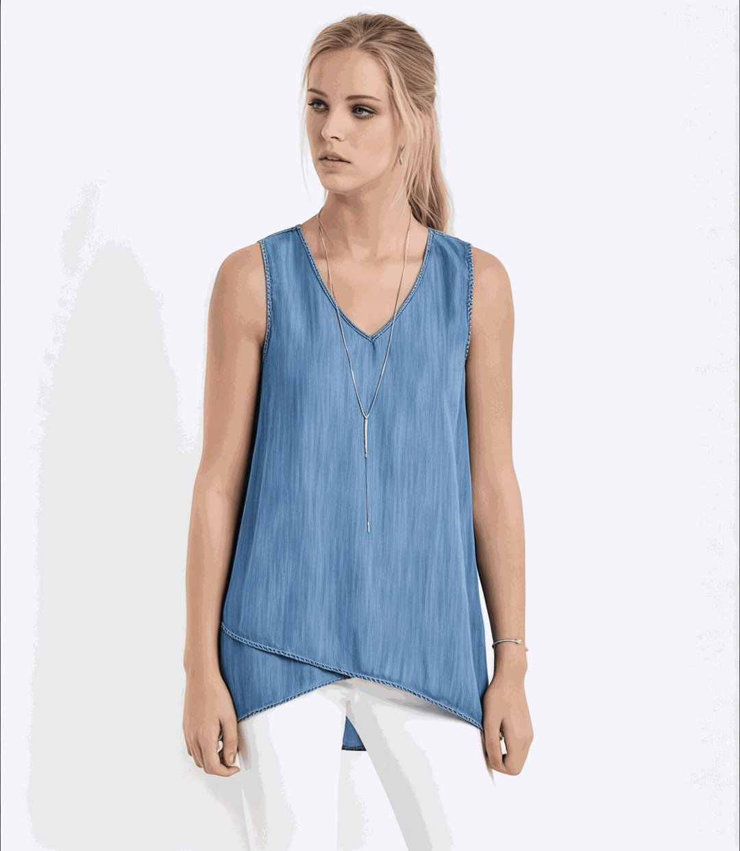 Woman wearing blue asymmetric tank top with crossover hem and V-neckline paired with white pants