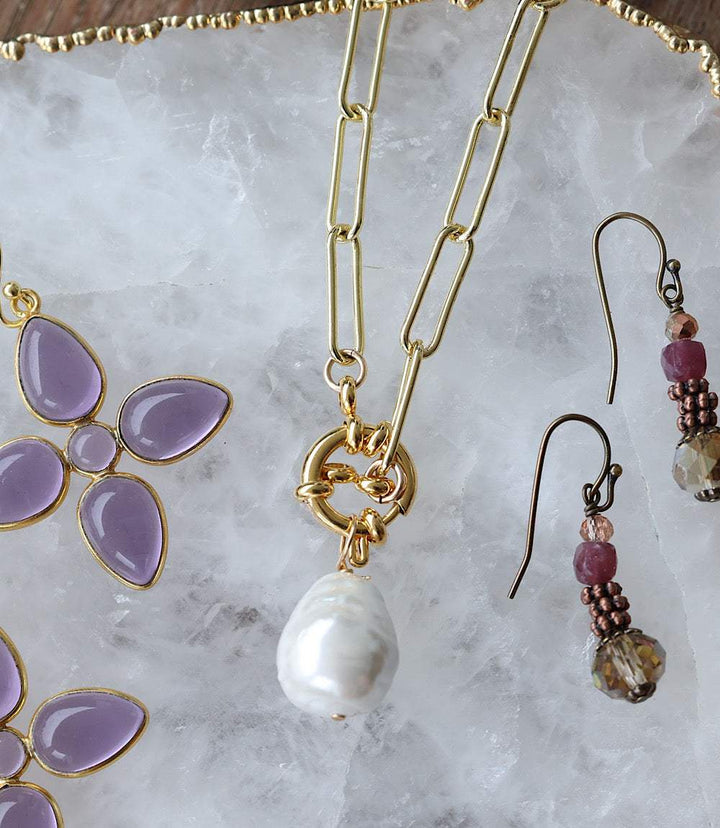Gold pearl toggle necklace with pearl drop on paperclip chain alongside purple flower earrings and dangling bead earrings on marble background