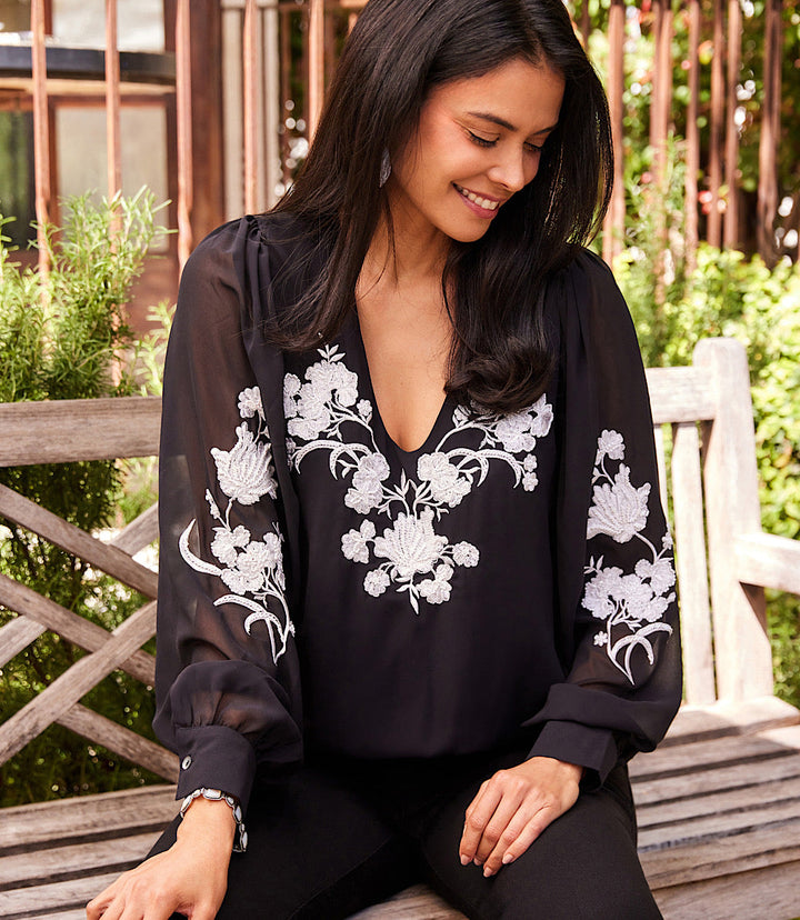 Woman wearing a black embellished blouson top with white floral embroidery and sheer sleeves sitting on a wooden bench outdoors