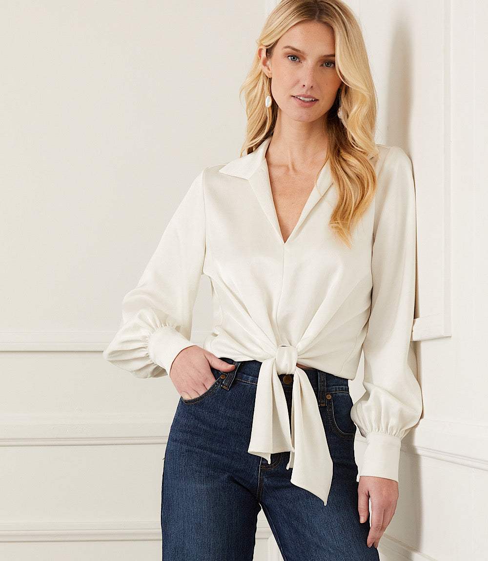 Woman wearing white satin tie front top with collared neckline and blouson sleeves paired with dark blue high-rise jeans