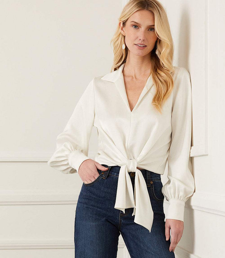 Woman wearing white satin tie front top with collared neckline and blouson sleeves paired with dark blue high-rise jeans