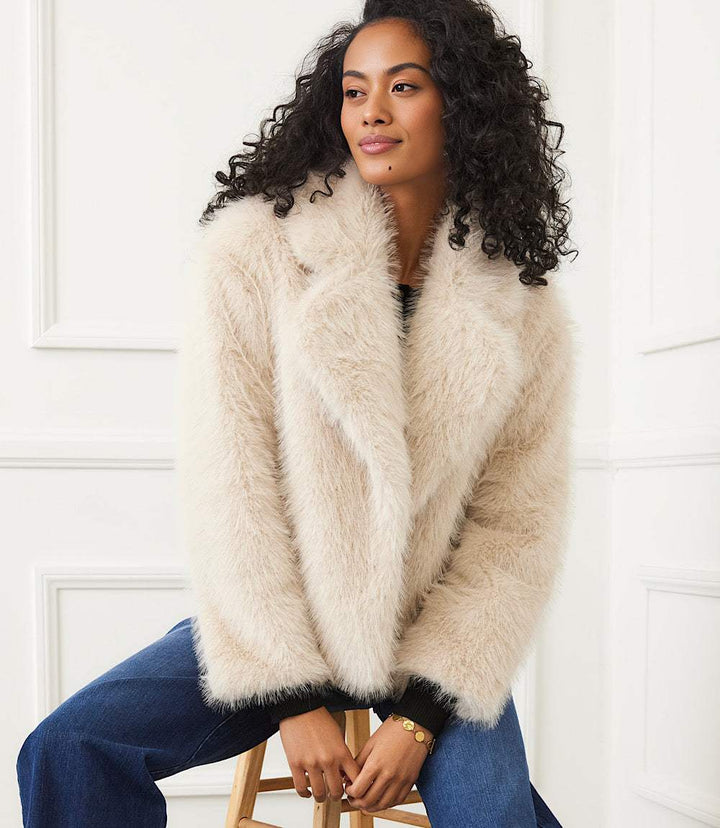 Woman wearing a cozy cropped faux fur jacket with wide notched collar and button-front closure sitting on a stool