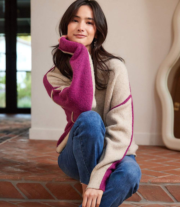 Woman wearing petite size colorblock mock neck sweater with purple and beige panels and dropped shoulder sleeves sitting indoors on brick floor