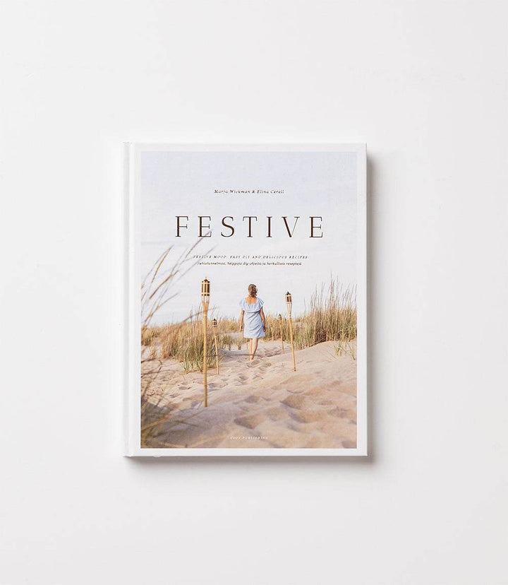 Cover of the book Festive: Life Is A Celebration featuring a person walking on a sandy beach with tall grass and tiki torches.