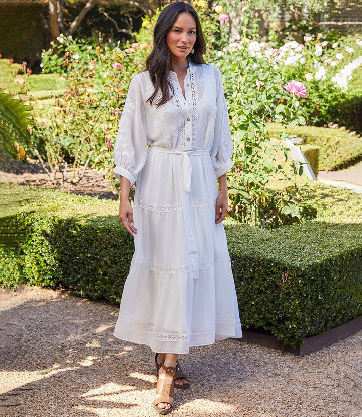 Woman wearing a white embroidered tiered dress with bishop sleeves and a tie belt in a garden setting