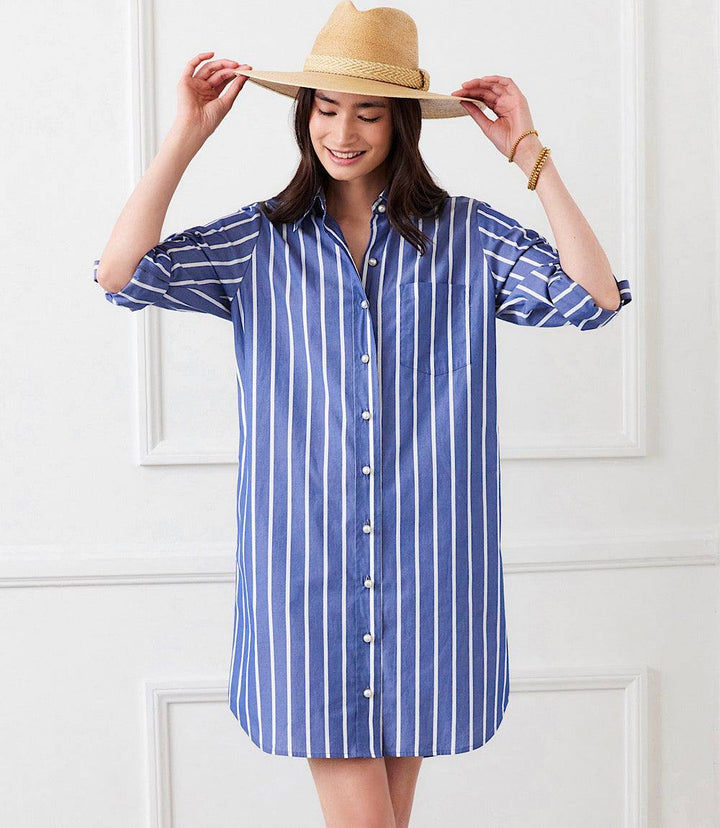 Woman wearing a blue and white Roll Tab Stripe Shirtdress with pearl buttons, collared neckline, and roll-tab sleeves, accessorized with a straw hat.
