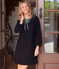 Woman wearing black puff sleeve embellished bow shift dress with scoop neckline and above-the-knee length