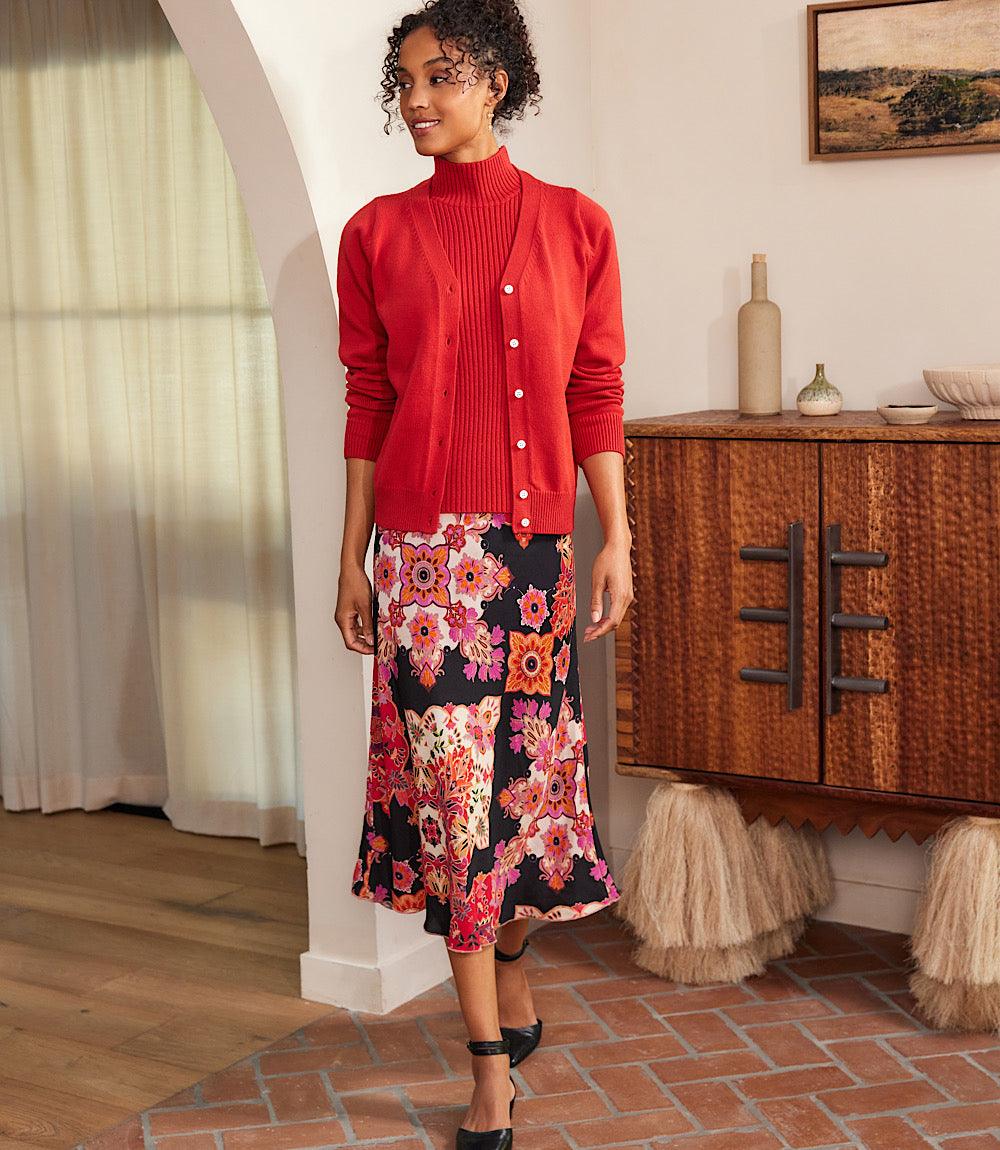 Woman wearing a petite size bias cut midi skirt with vibrant red, magenta, and cream floral print on black background paired with a red cardigan and turtleneck in a cozy indoor setting.