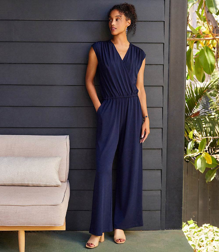 Woman wearing navy sleeveless travel jersey jumpsuit with crossover V-neck and wide legs, standing indoors with hands in side pockets