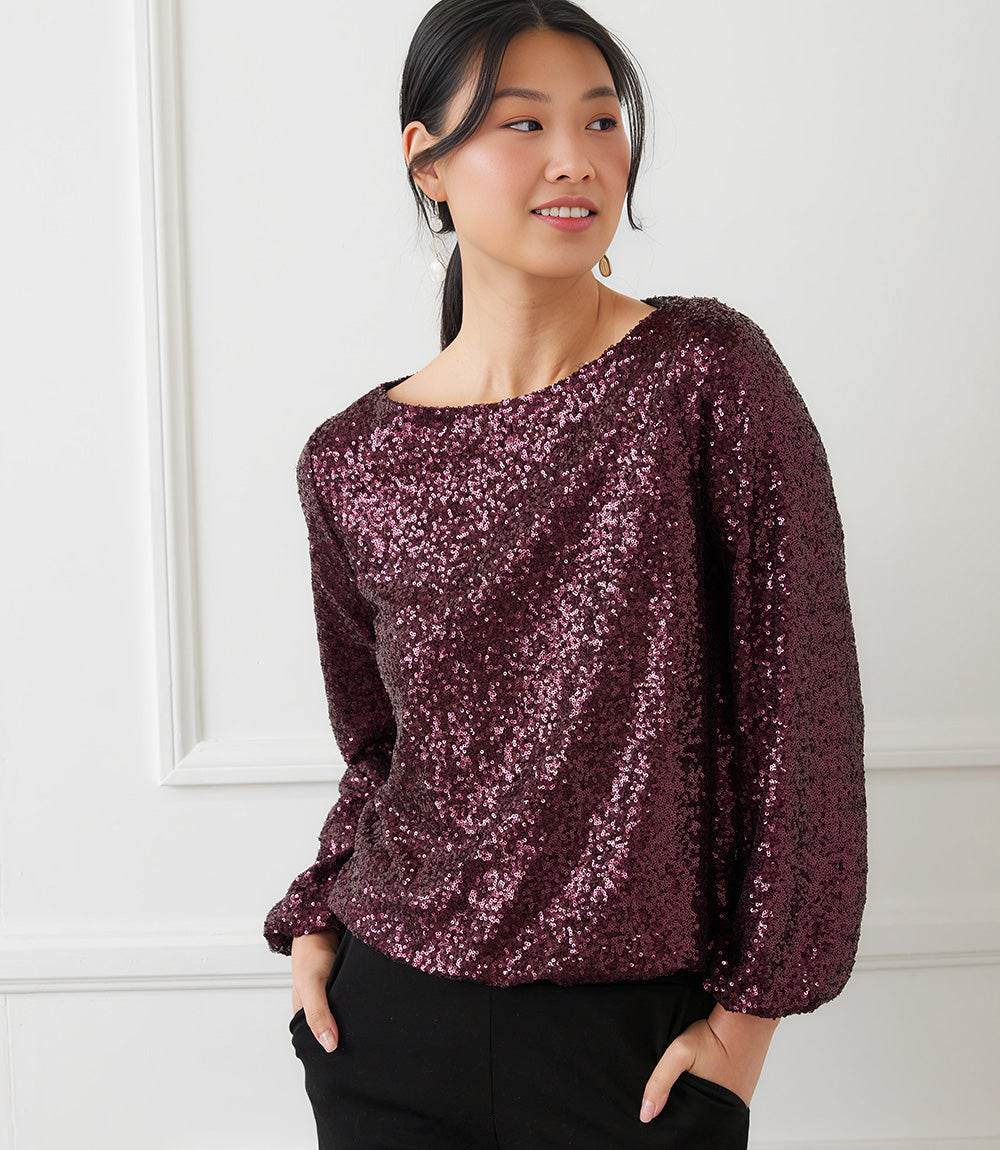Woman wearing a burgundy blouson sleeve sequin boatneck top with elastic cuffs and relaxed fit