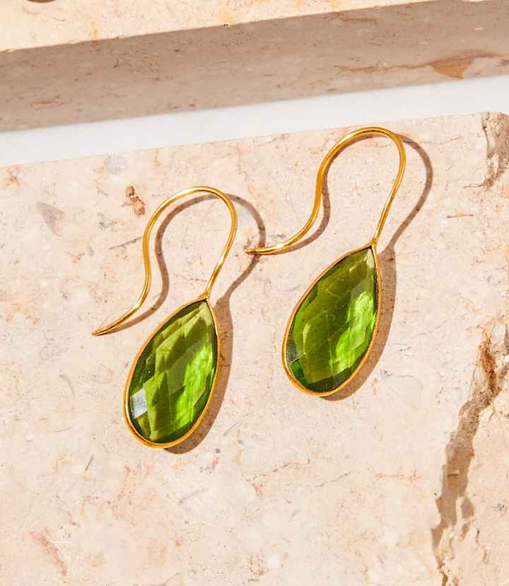 Long teardrop gemstone earrings with vibrant green peridot stones and gold plating on a textured stone surface