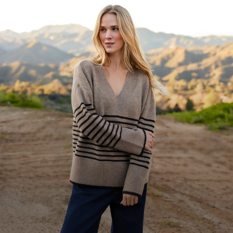 A woman stands outdoors on a dirt path with mountains in the background, wearing a taupe knit sweater with black horizontal stripes on the sleeves and chest, paired with dark blue pants.