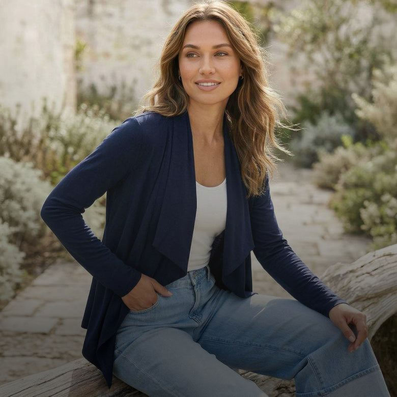 A woman sitting outdoors wearing a navy blue open-front cardigan over a white top and light blue jeans, with a blurred stone path and plants in the background.