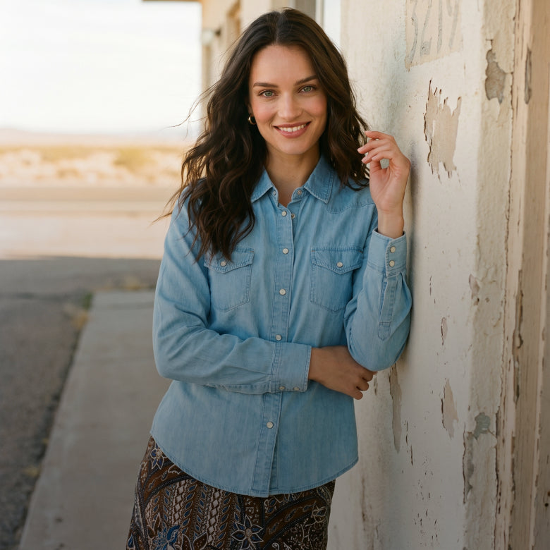 A woman wearing a light blue denim button-up shirt and a brown patterned skirt leans against a weathered white wall outdoors.