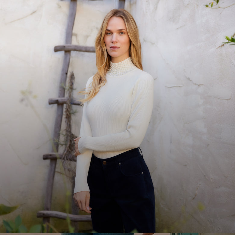 A woman wearing a cream mock neck knit sweater and dark navy pants, standing against a textured light beige wall with a rustic wooden ladder.