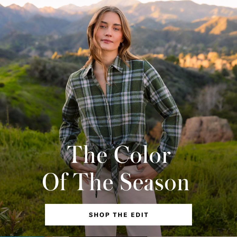 A woman stands outdoors in a mountain landscape wearing a green plaid button-up shirt tied at the waist with beige pants. Large overlaid text reads 'The Color Of The Season' with a 'SHOP THE EDIT' button below.