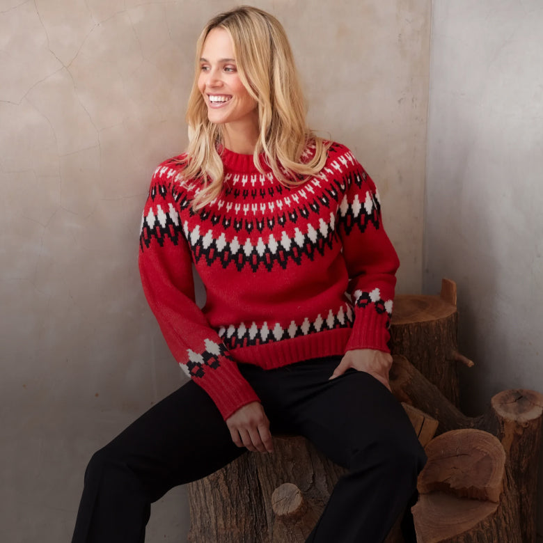 Woman wearing a red knit sweater with black and white geometric patterns, paired with black pants, seated on rustic wooden logs against a neutral stone wall.