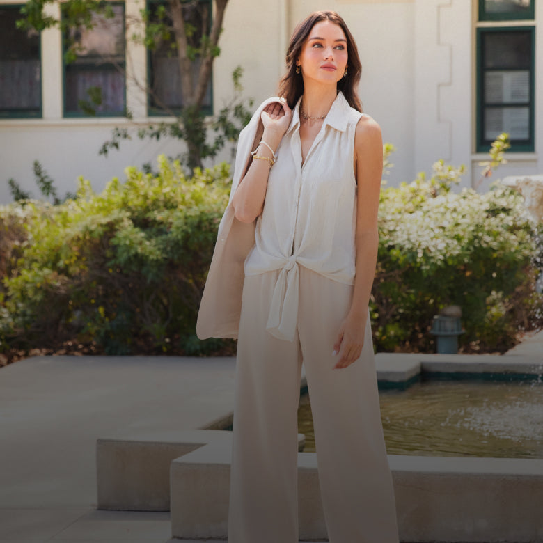 A woman stands outdoors in front of a building and garden fountain, wearing a sleeveless cream collared top with a front tie and matching wide-leg pants, with a light beige blazer draped over one shoulder.